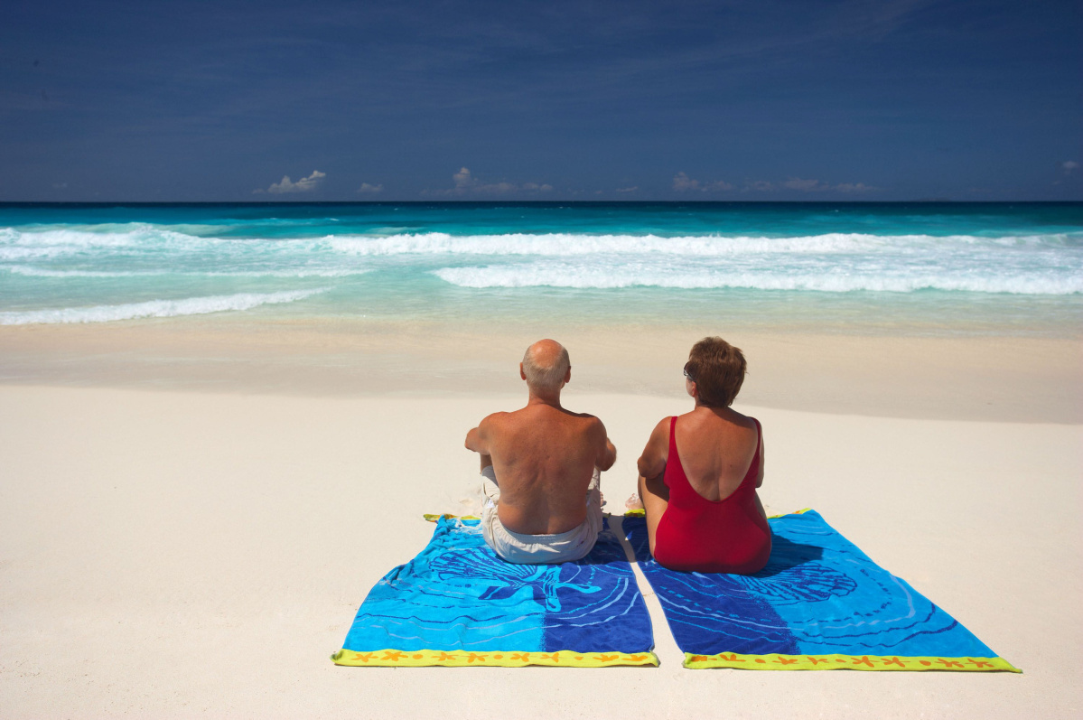 back view of senior couple sitting on beachtowels on sandy beach. Shutterstock